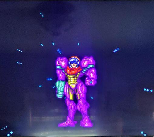 Samus Aran stands in her gravity suit against a mostly black void with stars shooting toward the foreground. She is sans helmet and showing a thumbs up.