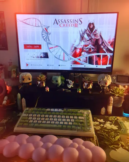 This is a picture of my gaming desk with pink and green accents and Assassin's Creed 2 on the monitor.