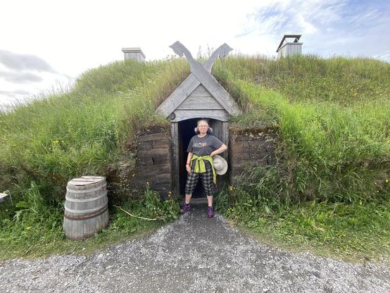 Me standing in the doorway of a sod longhouse. It has a grassy roof and there's a barrel to my side.
