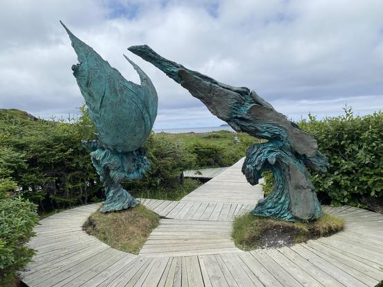 Abstract sculptures near the entrance to the site. They appear to be copper with a blue-green patina. They are craggy and windswept looking against a field of bright green with the ocean in the background.