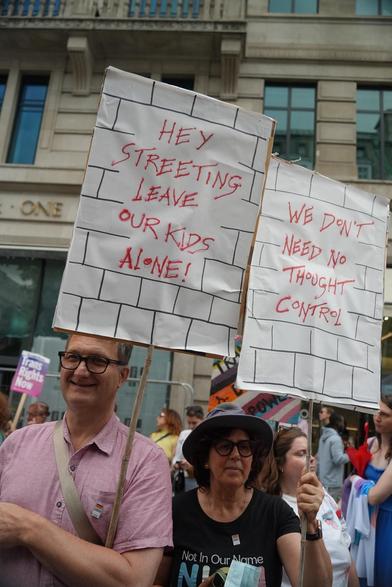 Two people holding matching protest signs with text over a hand drawn brick wall texture. The left sign says "Hey Streeting Leave Our Kids Alone!" The right one says "We don't need no thought control!