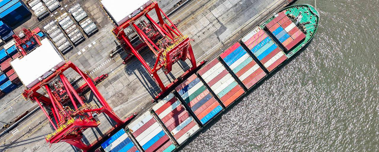 Frachter in Vogelpespektive. Container, die hauptsächlich in weiß, rot- und blau-Tönen gehalten sind, sind in 9er Reihen auf dem Schiff geordnet und erinnern dabei ein wenig an die Trans Flagge bzw an die Climate Stripes.