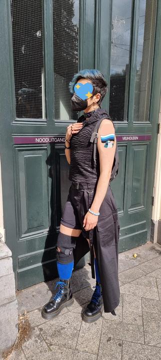 domo standing outside blocking an emergency exit to a building. They're wearing the same outfit from the previous post, all black goth vibes. Black sleeveless shirt, black shorts, black semi-skirt tied around the back, a black knee brace, blue socks, black platform docs with blue laces, and a black backpack. They also have black rings, bracelets, choker, beaded necklace and a blue and white beaded bracelet that says "he/they". You can also see they have on a black FFP2 mask and they have a quake Q earring that has a chain that connects to their industrial piercing. They have one hand over their heart. The photo is very sunny.