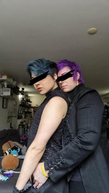 scremlin and domo in a living room posing for the camera. Domo has blue hair and scremlin has purple hair. They both have black scribbled out eyes. Scremlin is holding domo from behind. They are a power couple.