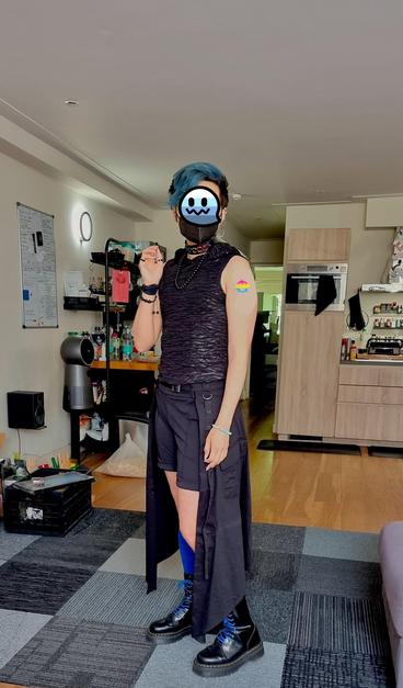 Full body half turned photo of Domo, a pale latinx person, in a living room wearing black shorts, a black tie around half long skirt, calf high dark blue socks, black platform docs with blue laces, a black FFP2 mask, and a black choker. They have have several black bracelets and a black beaded necklace. Their eyes have been blocked out with a transmasc ghost emoji and their arm has a pansexual flag colored anarachy symbol.