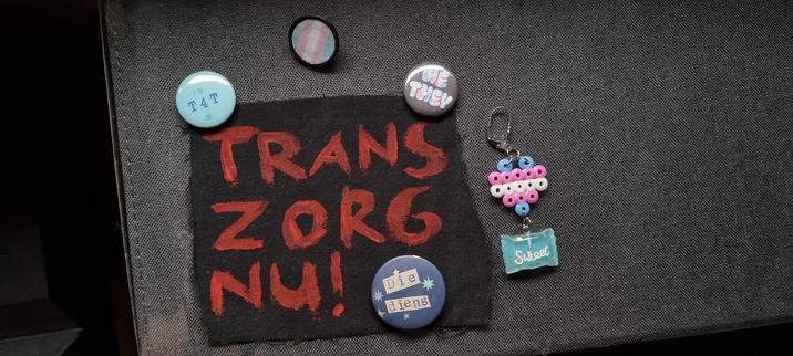 Photo of a large scrap of black fabric that says in red fabric paint "Trans Zorg Nu!" (Trans healthcare now!) to be used a patch. Alongside the patch are 3 small buttons that say "T4T", "He They", and "Die Diens" (they/them in dutch) respectively. There's also a small round trans flag pin and a trans flag heart earring that has a dangly bit that looks like a wrapped teal candy with white letters that say "sweet".