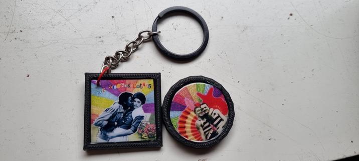 Photo of two queer art pieces . On is on a keychain and it looks like a framed black and white photo of two Black masc people kissing with the text "We always existed" above them and a rainbow background. The second is a magent with a black and white photo cut out of two white men holding eachother close in swimming suits with rainbow colored background, also framed in black.