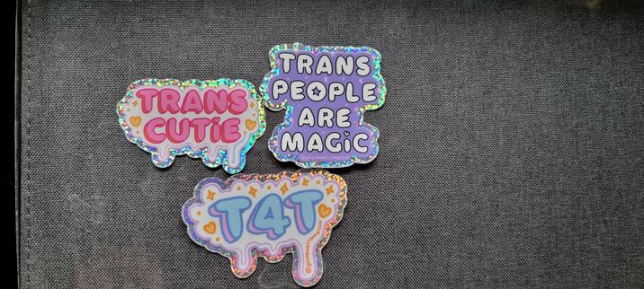 Photo of three stickers in very cute bubble letter font with sparkly edges.

Sticker 1 says "Trans Cutie" in pink text on light link background with orange sparkles and hearts.

Sticker 2 says "Trans people are magic" in white text on purple.

Sticker three says "T4T" in blue with pink shadow on white with orange sparkles and hearts.