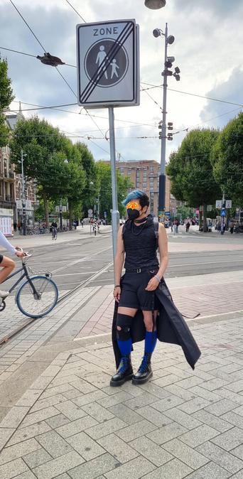 Full body photo of Domo in the same outfit in Amsterdam outside during the day. They're standing next to a no walking with children sign on the sidewalk. You can see their skirt thing flow in the wind.