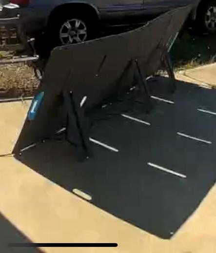 A portable folding photovoltaic panel is placed on a concrete driveway, with a vehicle partially visible in the background. The panel is dark with three support legs, casting a shadow on the ground.