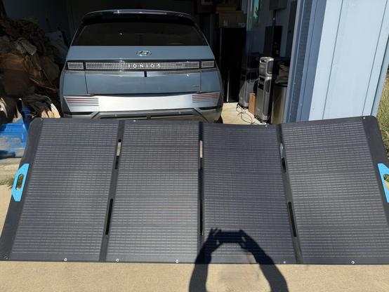 A portable solar panel is positioned in the foreground, with a Hyundai Ioniq 5 electric vehicle visible in a garage. The F3800 batteries and home power panel are visible along the wall inside the garage. Thin black cables from the back of the PV panel are along the floor of the garage.