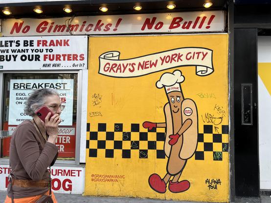 Photo by Fenichel:

A street-corner scene in front of "Gray's Papaya", on New York's west side.
A woman walks into the frame from the left side, behind her a white panel with a sign: "LET'S BE FRANK... WE WANT YOU TO BUY OUR FURTERS...
HEH HEH.... GET IT?" [The words FRANK and FURTERS are in red]

A sign at the top, across the wall says; "No Gimmicks! No Bull".
In the main part of the frame (2/3 of the right part) is a yellow background and a bit of art-deco-like street art:  A frankfurter, with white chef's hat labeled "MR FRANK", and big red shoes, his body being a hot dog and bun, is standing at a checkerboard pattern, a banner overhead announcing "GRAY'S NEW YORK CITY"