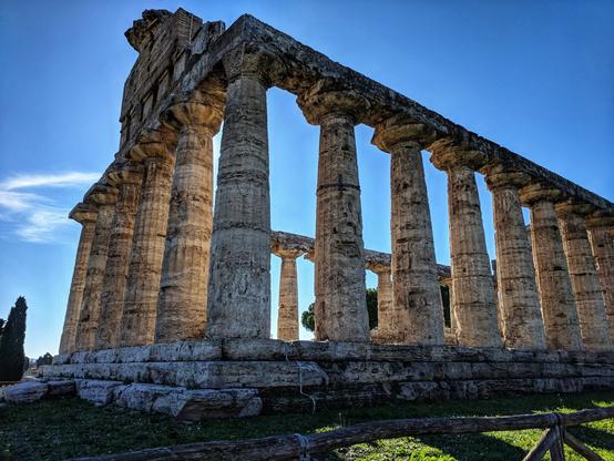 A low-angle shot of the partially ruined Temple of Athena at Paestum, Italy. The massive, weathered Doric columns are bathed in sunlight against a clear blue sky, casting long shadows. A few stone blocks lie on the ground at the base of the temple. In the foreground, a simple wooden fence runs along a patch of green grass. A small part of the temple's roof structure remains on the left side, showing intricate stonework.