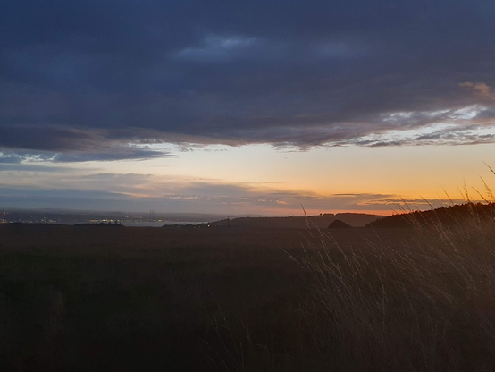 Photo of a landscape. In the foreground of the photo is some tall pale yellow grass and the expanse of a field. To the left of the photo, in the distance is the river Tay, the Tay road bridge and the lights of Dundee. An oil rig is in at the docks of Dundee, its tall towers add some shape to that side of the river. To the right are the gentle hills of Fife. The sky is dominated by a large dark cloud, but the distant sky is lighter, with dawn breaking, gentle yellows and pink colour the sky.