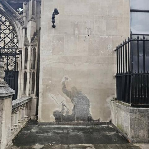 A view from the street, showing one entrance to the Royal Courts of Justice in London, off to the left side; the center of the photograph is the wall where a Banksy has been erased, leaving behind the black and white silhouette of a bewigged judge, holding a gavel like a hammer over their head while standing over a prostrate protester, who is unarmed, holding up one empty hand and a placard on the other hand. The stain is very clear against the rest of the wall.