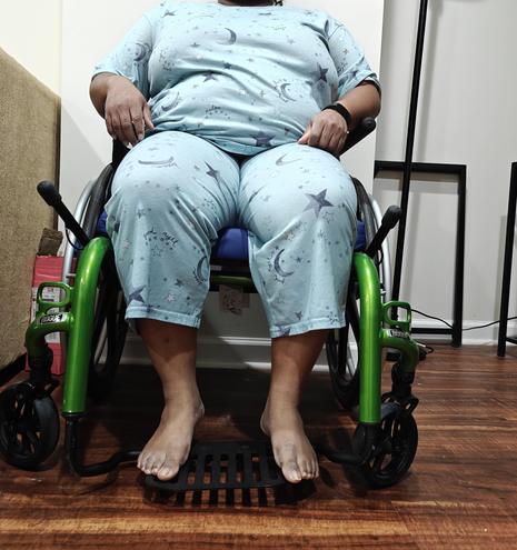 A Black person in blue pajamas with black stars on them sitting in a green wheelchair. They're struggling to keep their feet on the foot plate