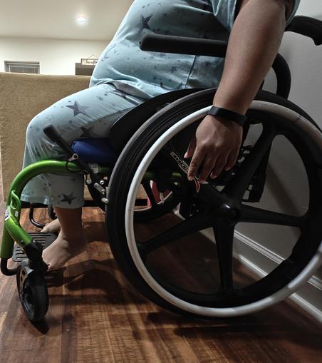 A side view of a black person in blue pajamas with stars on them sitting in a green and black wheelchair. Their hands do not reach the center of the wheel