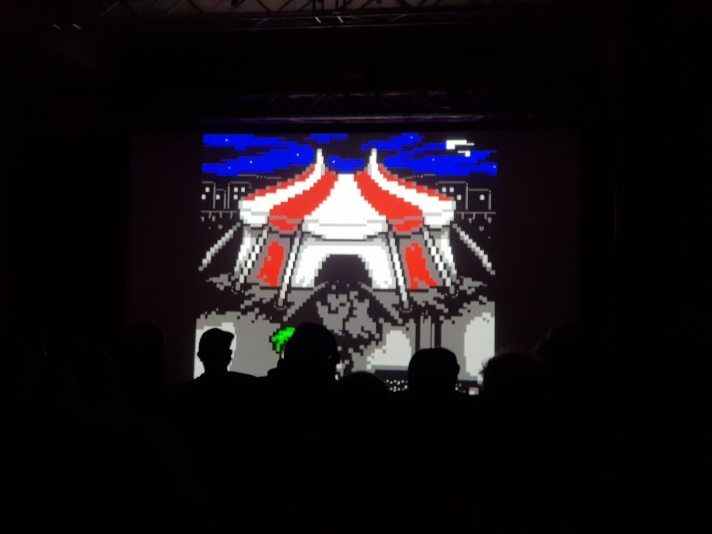 A photo of the opening circus tent in "Circus of Dreams" shown on the projector screen at Deadline 2024, with the silhouettes of the demoparty audience visible in front of it.