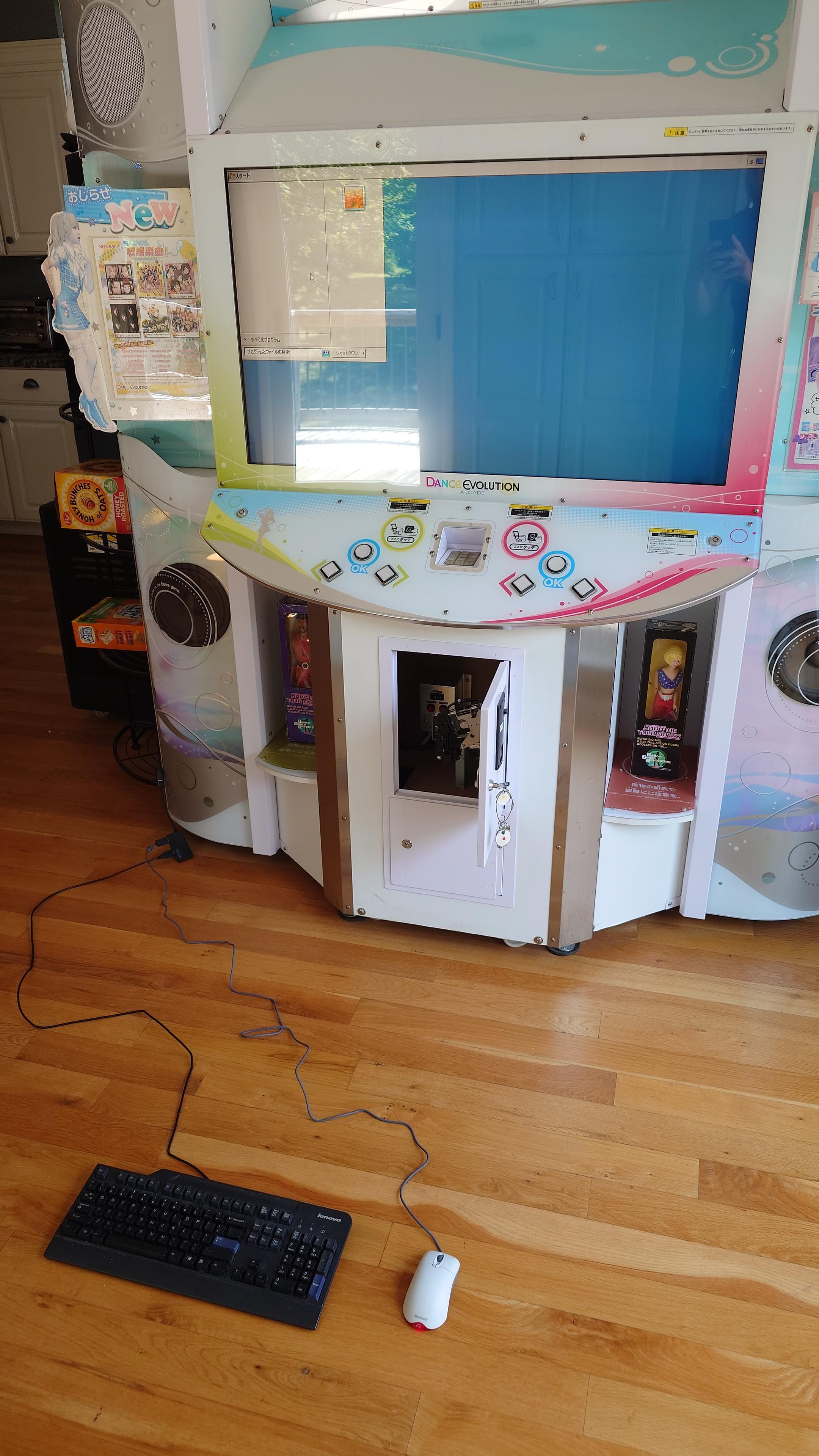 A dance evolution arcade cabinet showing the Windows 7 embedded operating system without the game running. On the floor in front of it are a Microsoft intellimouse optical mouse and an old Lenovo keyboard, plugged in via a USB hub that trails off to the side of the cabinet.